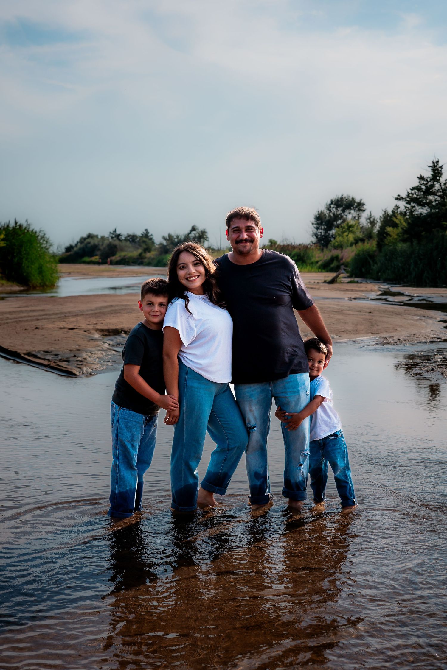 Happy family of four standing barefoot in shallow water at a sandy beach, smiling together on a cloudy day.