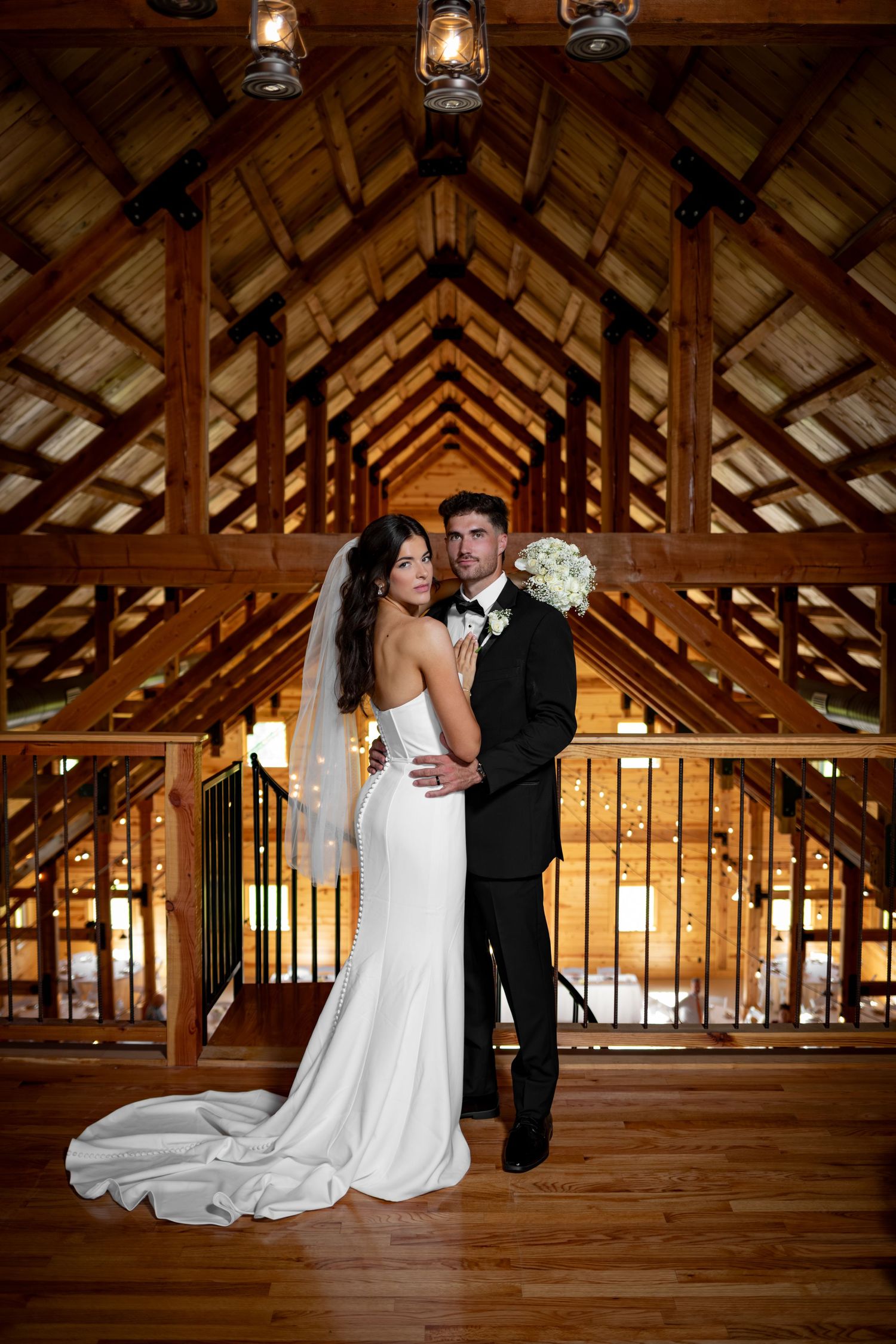 Bride in white gown and groom in black tuxedo pose in rustic wooden barn venue with vaulted ceiling.