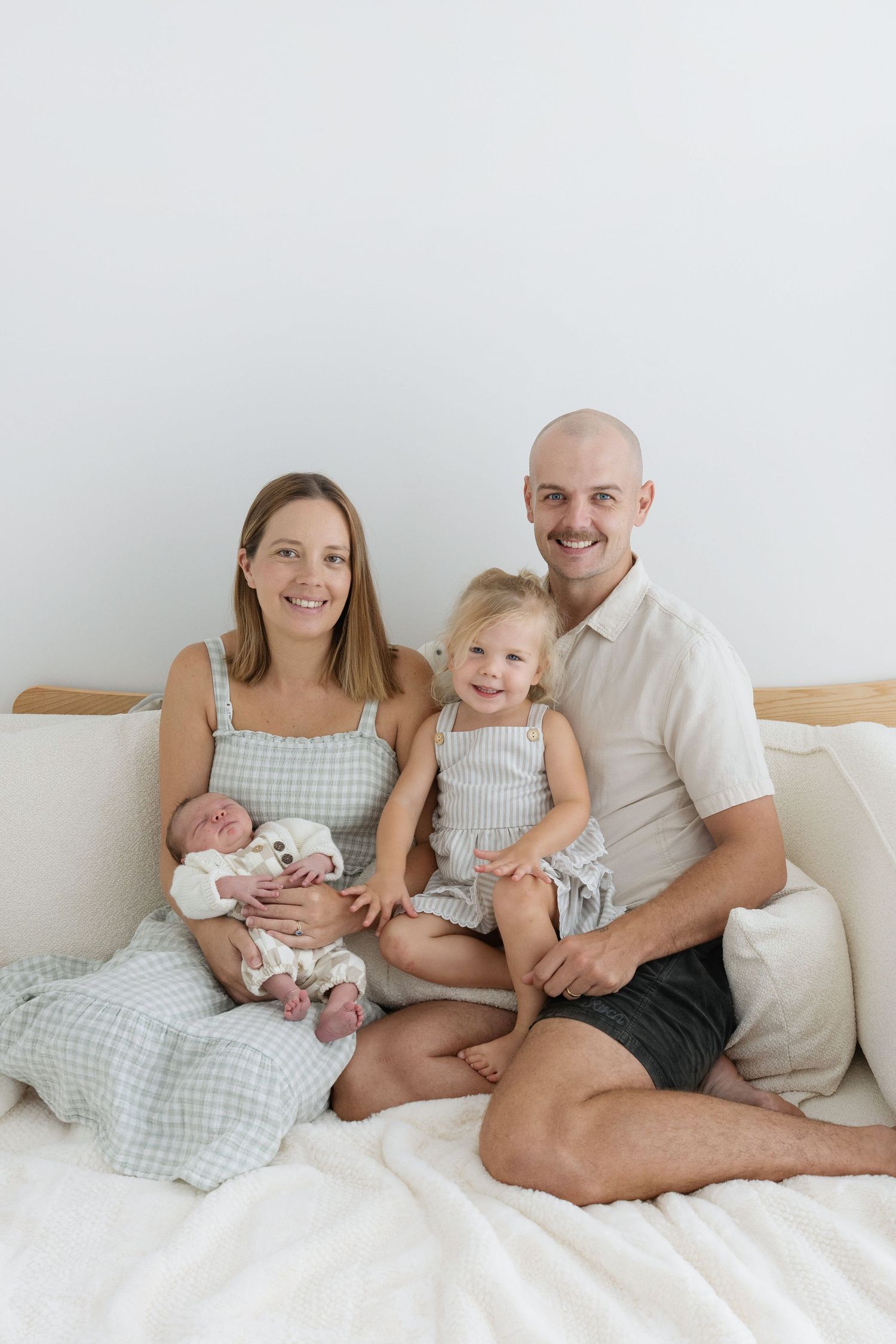 Happy family of four sitting on a white bed, mother holding newborn baby, toddler smiling, father embracing children.