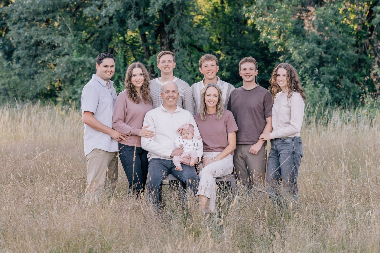 Large family group portrait in a golden meadow with lush green trees, featuring adults and a baby in warm tones.