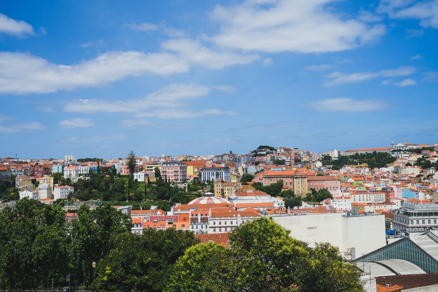 Panoramic view of Lisbon cityscape with terracotta rooftops, lush green trees, and a hilltop castle under a blue sky.