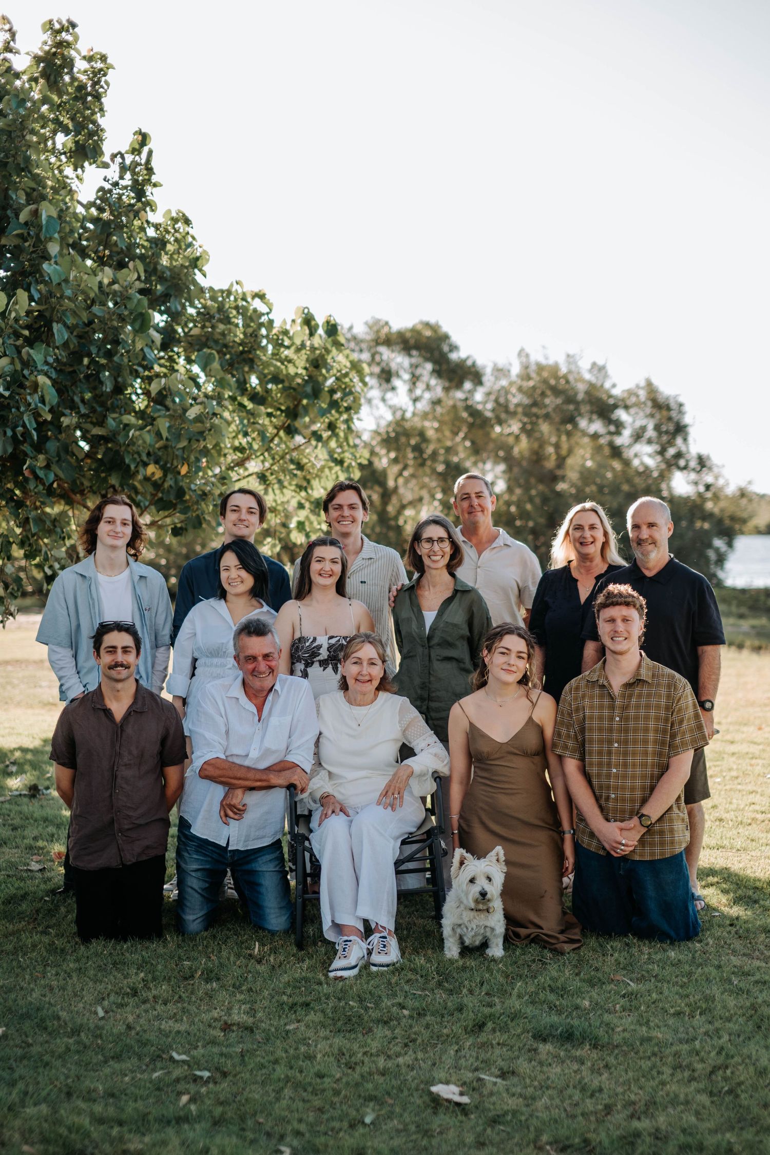 Large family group portrait outdoors on green grass with trees, including a small white dog, posing for a formal photo.