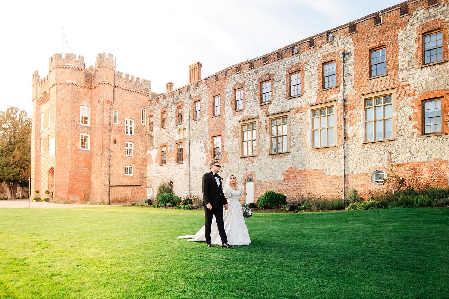 Bride and groom stand on lush green lawn before historic brick castle wedding venue on sunny day.