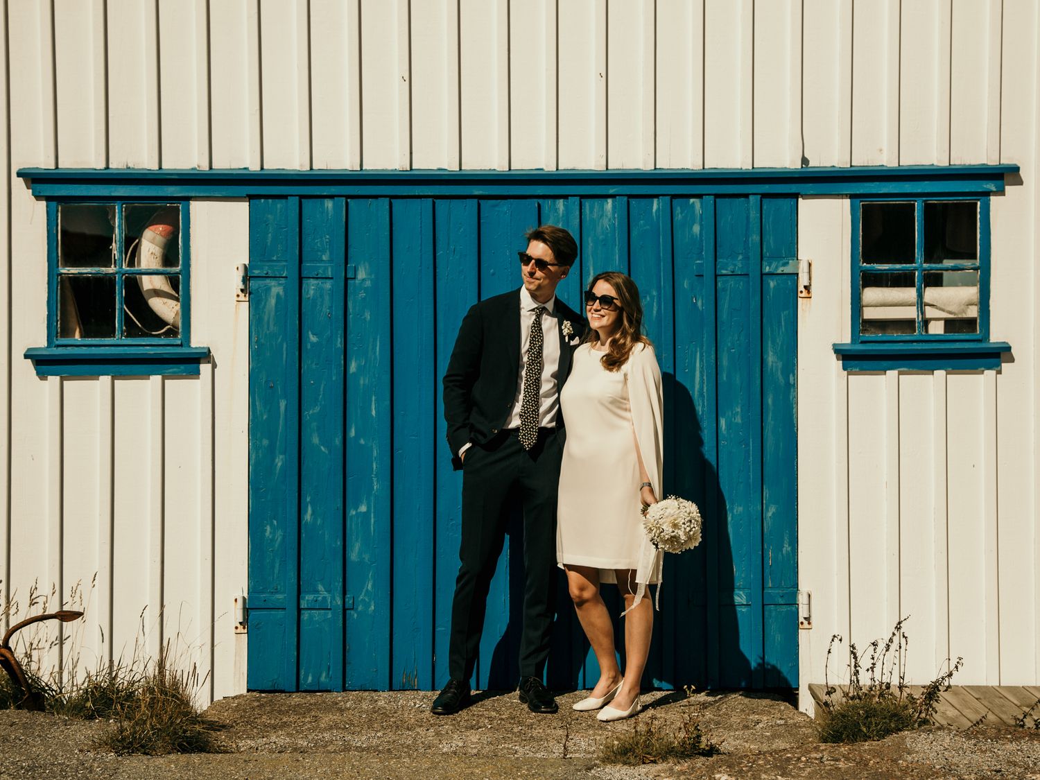 A couple poses against a rustic white building with bright turquoise blue doors and windows on a sunny day.