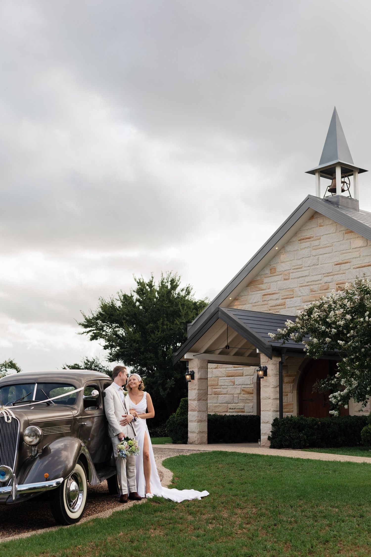 Vintage brown Chevrolet car parked outside stone chapel with cloudy sky backdrop on wedding day.