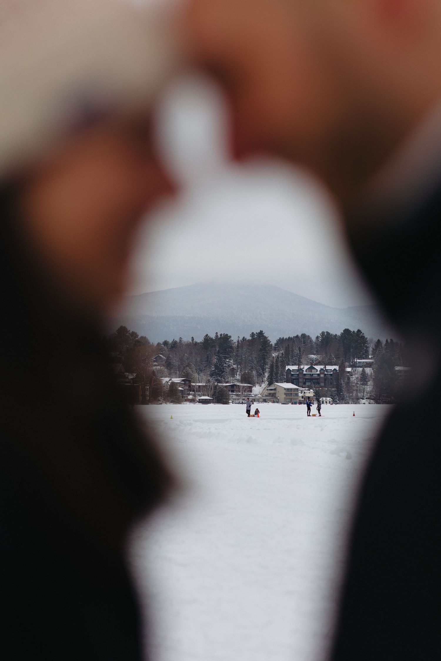 Through a blurred foreground, people can be seen on a snowy winter landscape with trees and buildings in the distance.