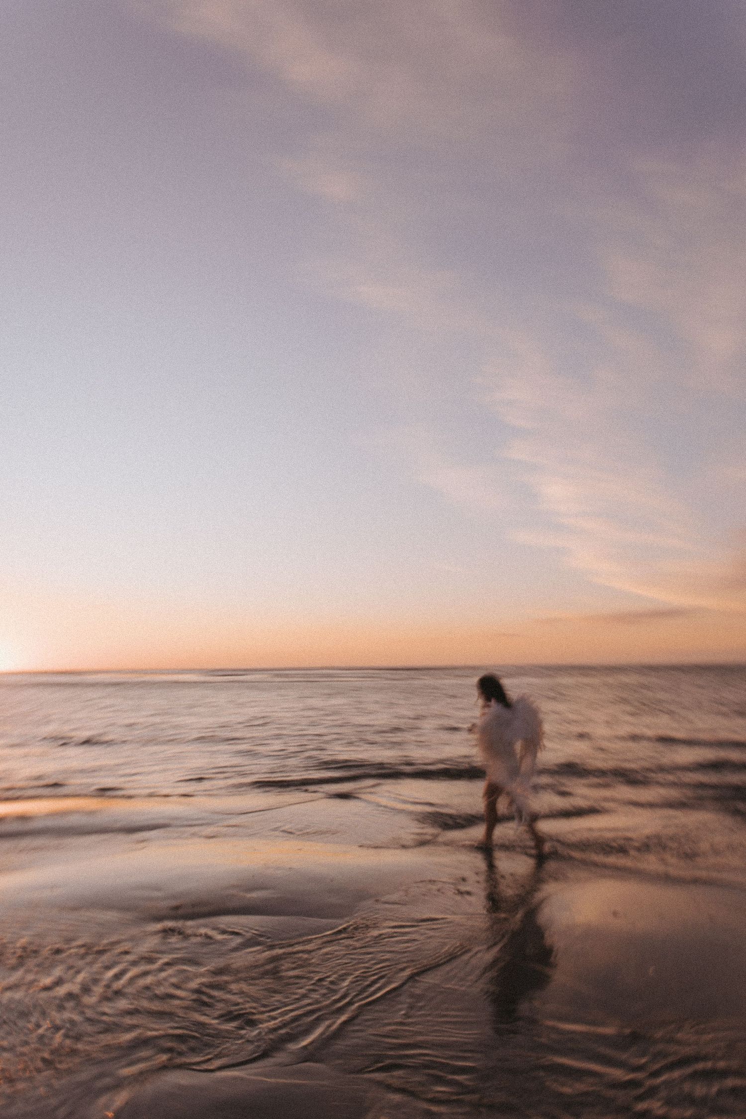 A woman in white stands on a sandy beach with arms raised, under a dramatic cloudy sky near the ocean.