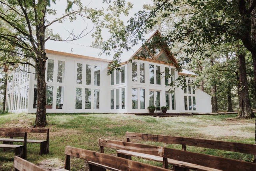 Historic white church building with tall windows and wooden benches sits among trees in a peaceful rural setting.