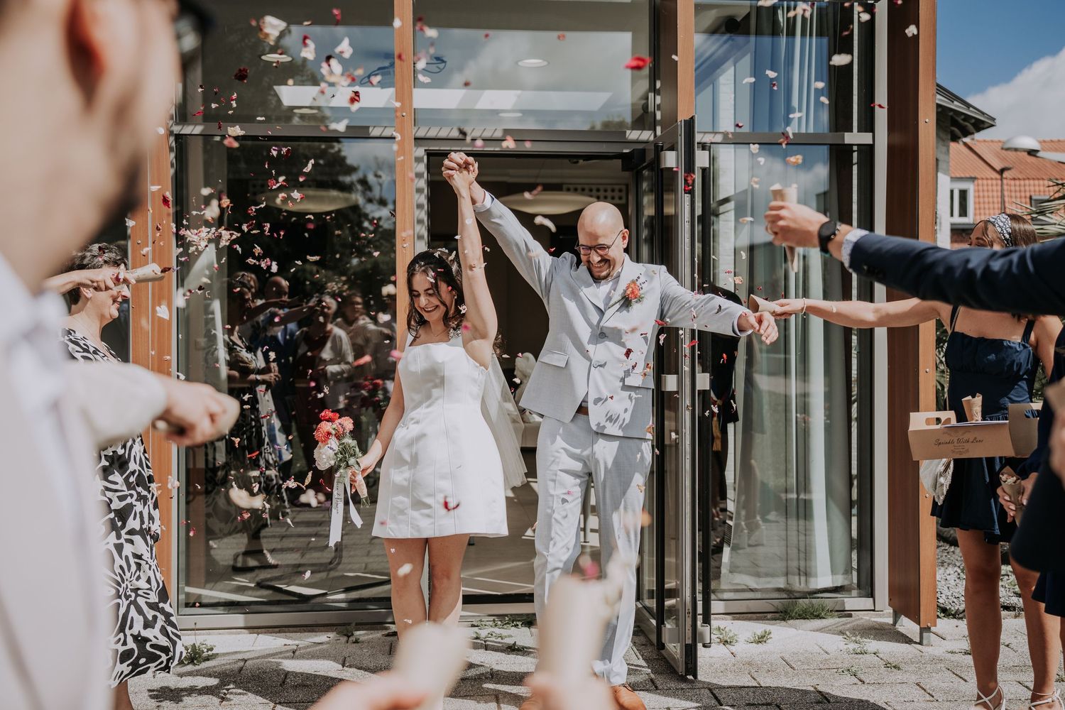 Bride and groom exit wedding venue celebrating with confetti thrown by guests on a sunny day.