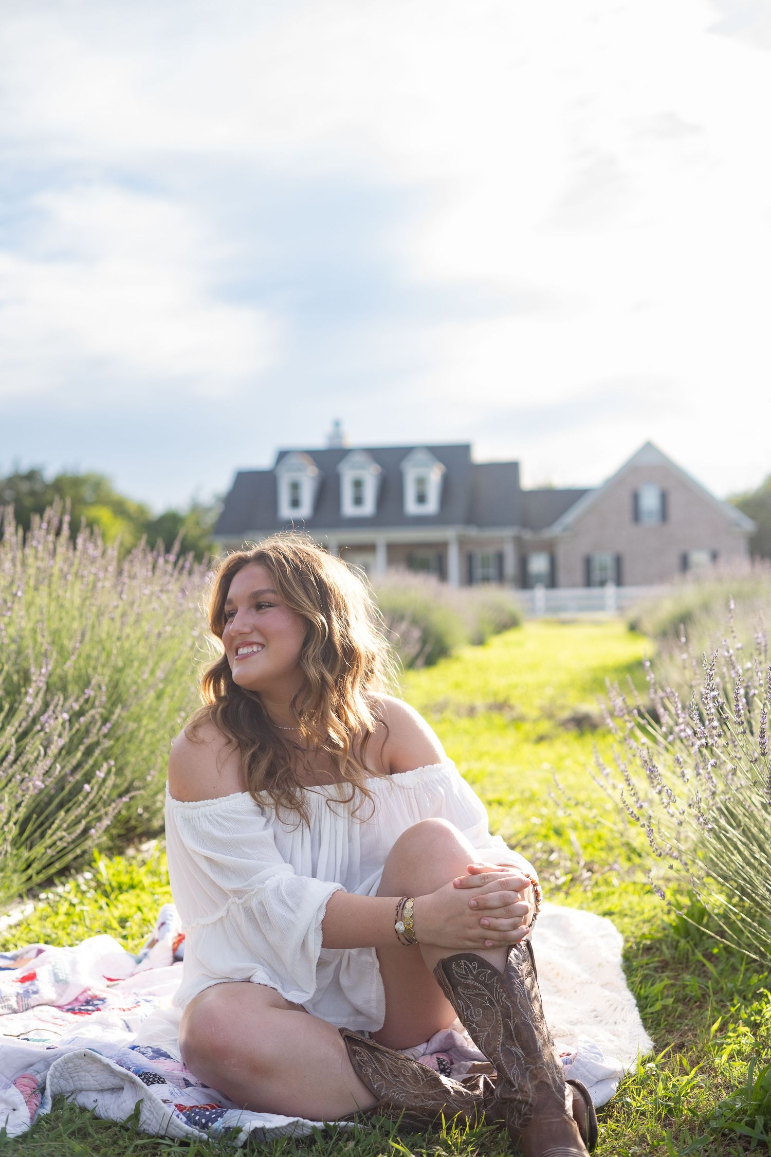 Young woman in white off-shoulder dress and cowboy boots sitting on blanket in lavender field near farmhouse.