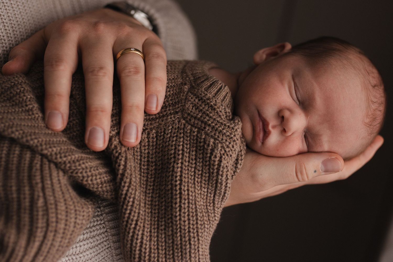 Newborn baby sleeping peacefully in parent's hands, with natural daylight highlighting tiny features. Image is in soft natural earthy tones