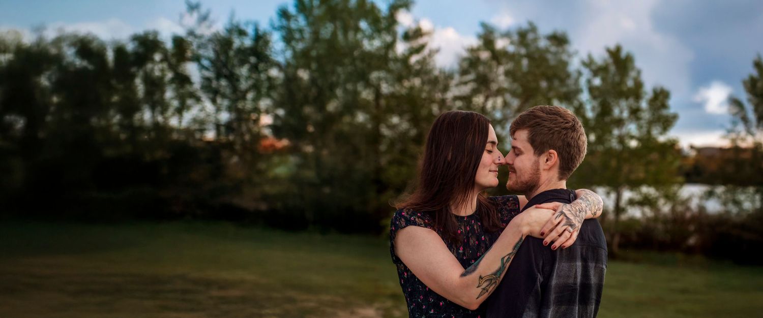 A Kentville Nova Scotia Mom & Dad embraces outdoors at dusk, surrounded by lush green trees and warm golden light.