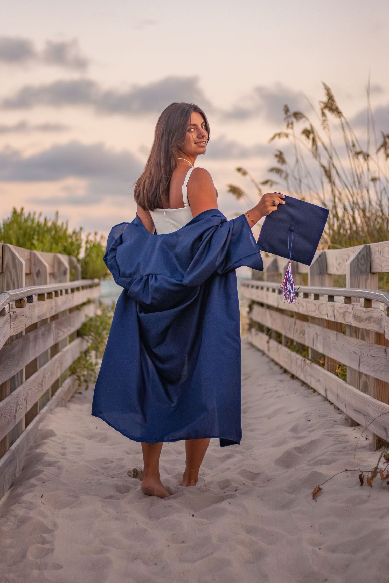 Graduate in navy blue gown stands barefoot on sandy beach boardwalk at sunset with sea grass in background. Senior Photos Graduation Photos