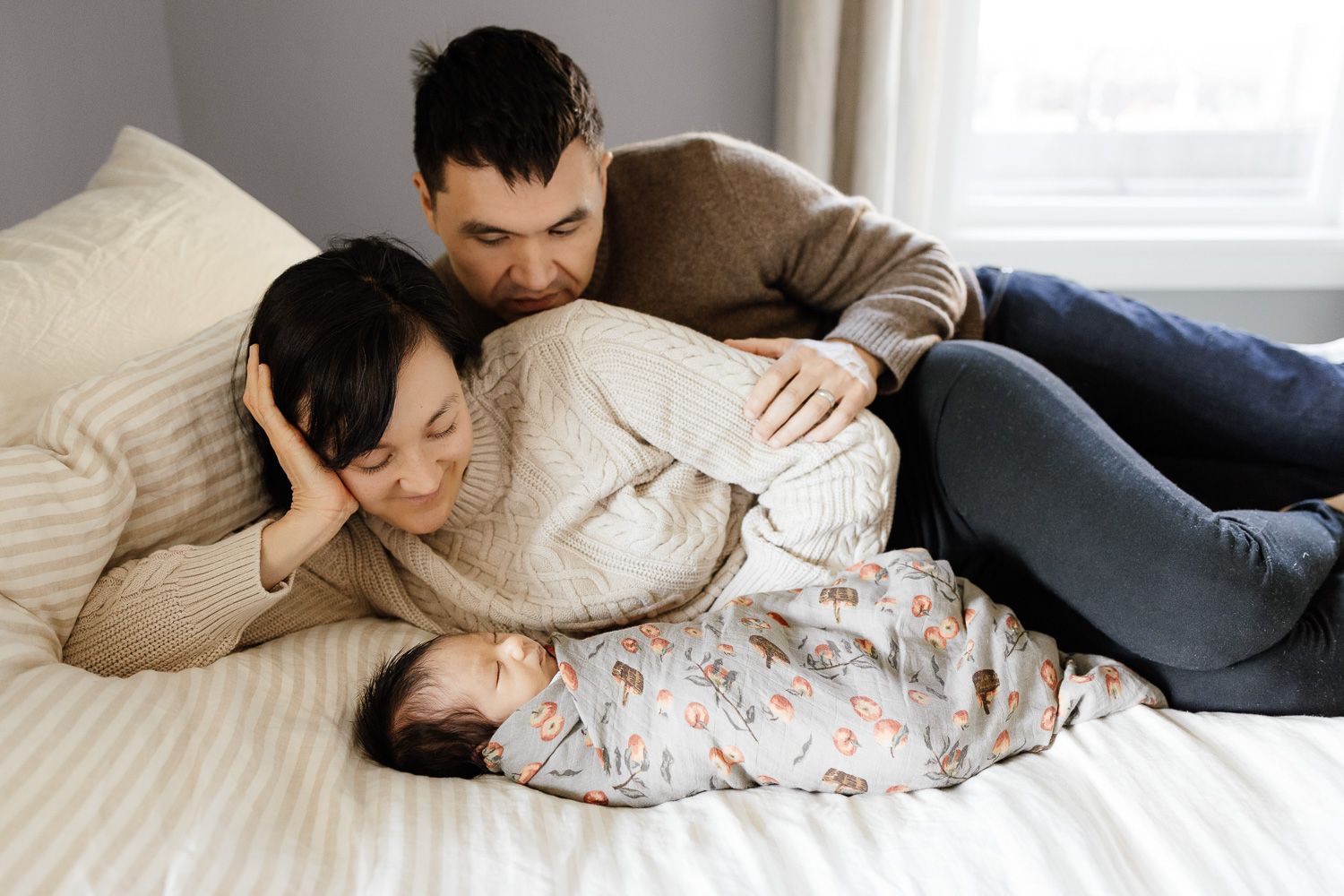 A couple rests together on a white bed with their 4 week old baby sleeping by them