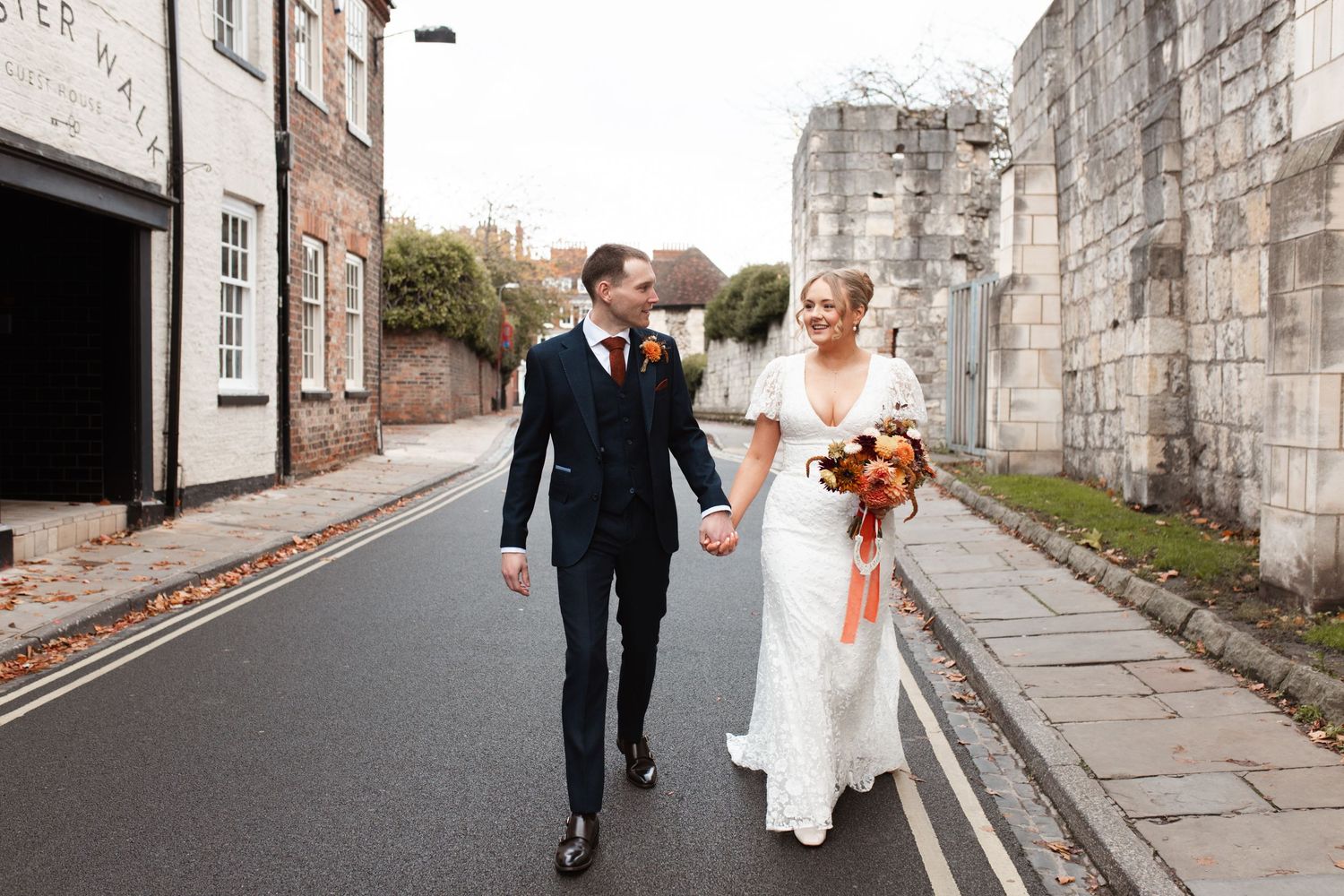 A couple walks along a historic stone wall in formal wedding attire. York Registry Office Wedding Photographer. York Museum Gardens.