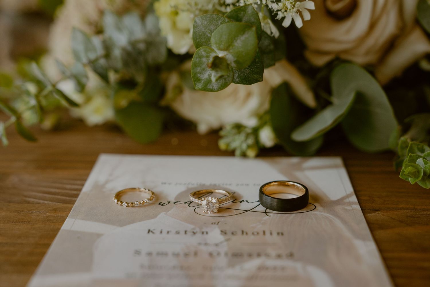 Wedding rings and invitation displayed on wooden table with soft floral arrangement in background.