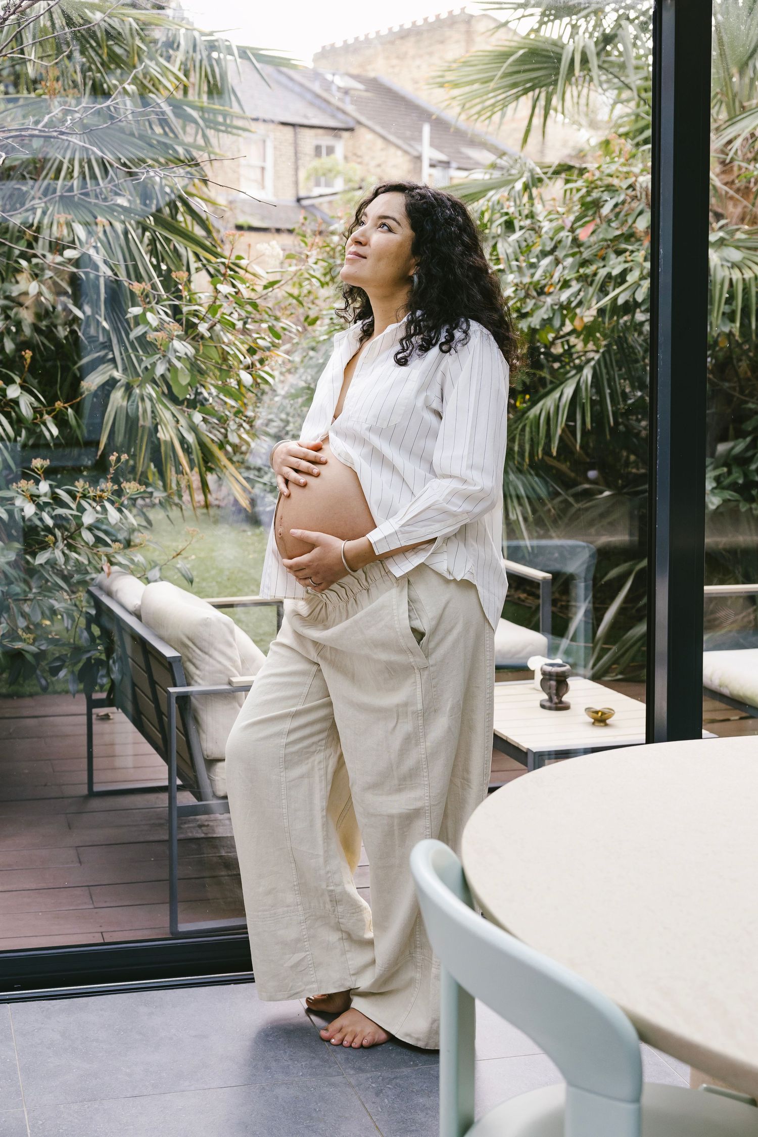 A person in an all-white outfit relaxes in a bright sunlit room with tropical plants visible through large windows.