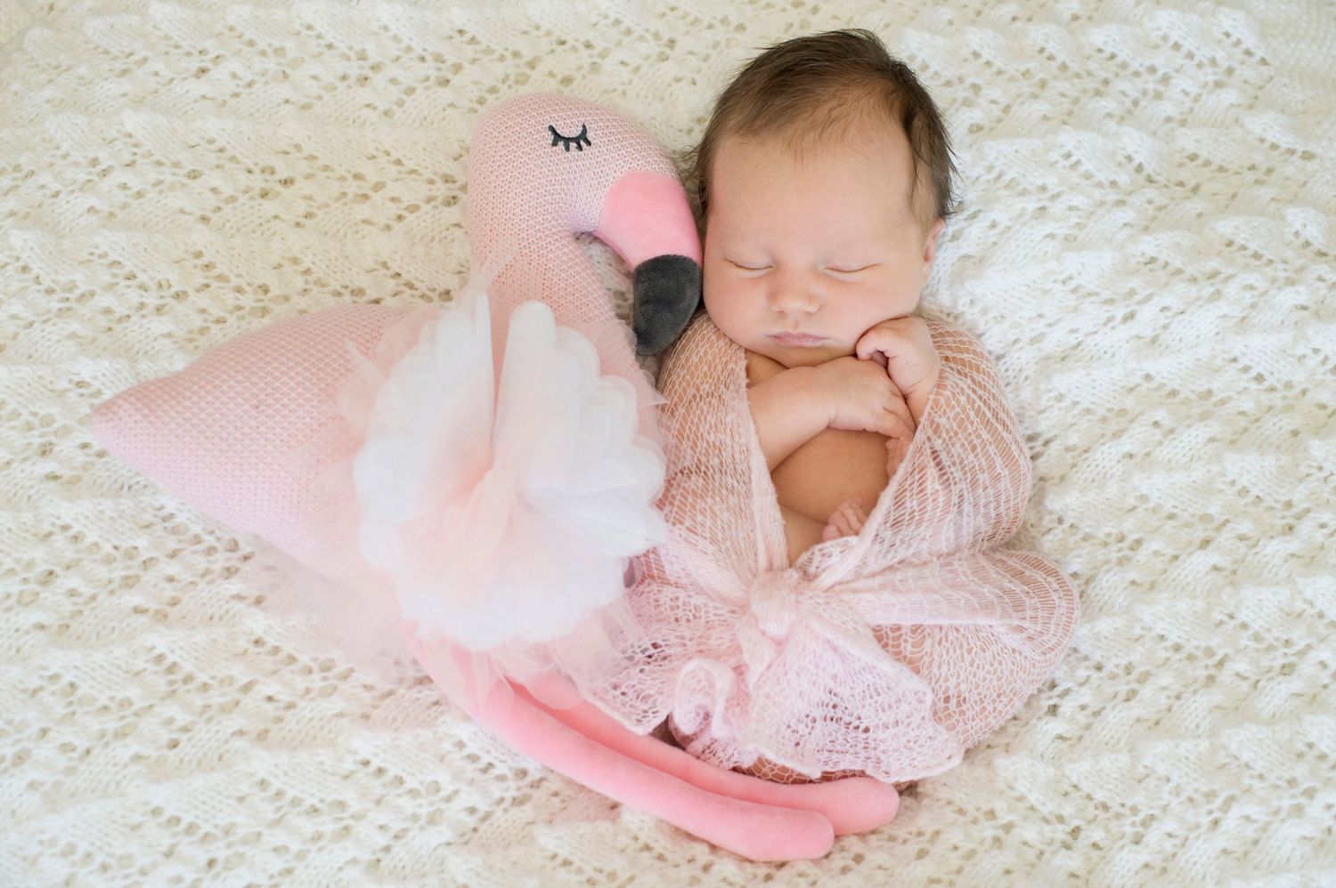 Newborn baby sleeping peacefully wrapped in pink knit beside a pink flamingo stuffed animal on a white blanket.