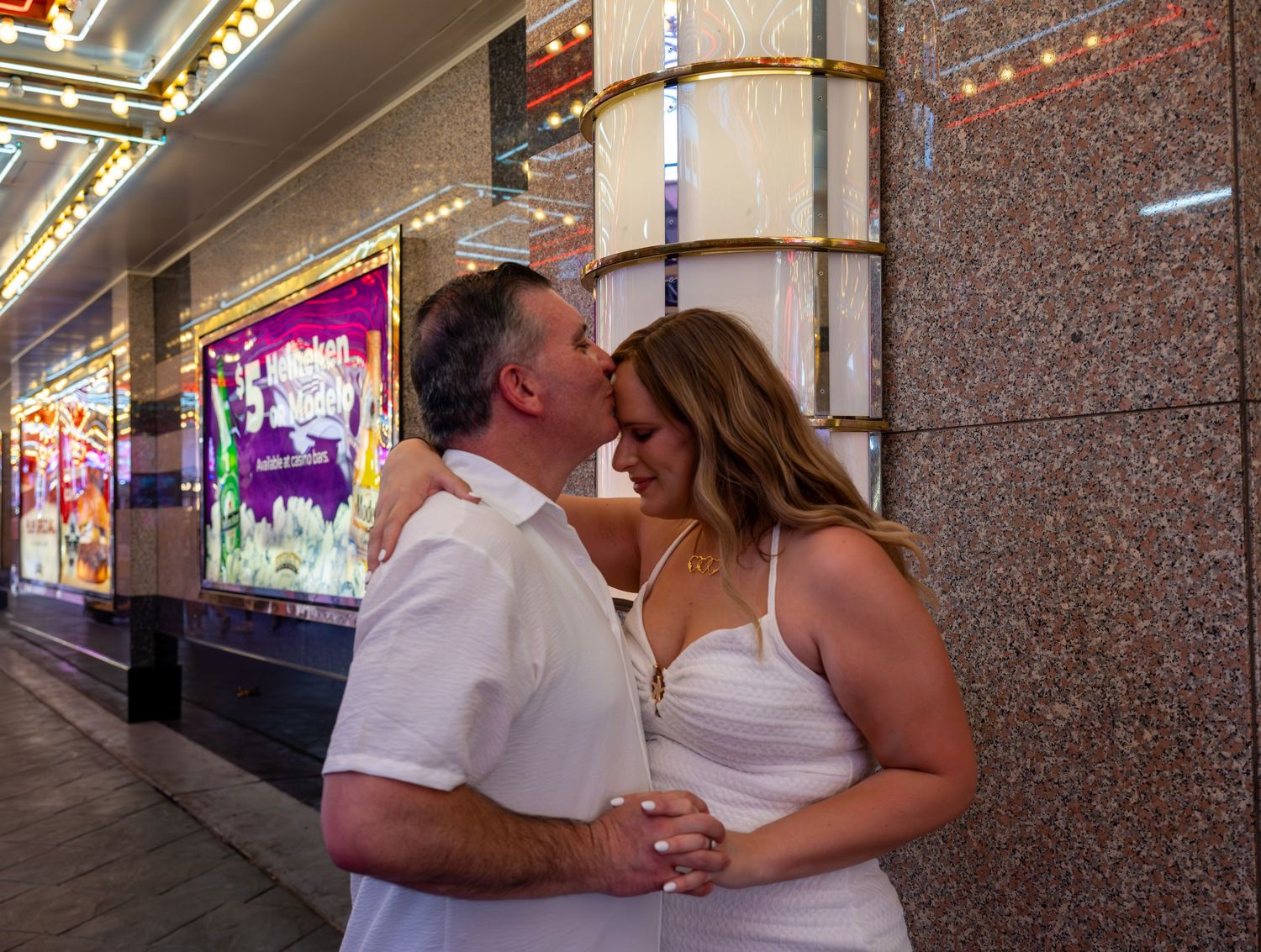 Couple sharing a romantic moment outside a bright theater marquee at night, dressed in white.