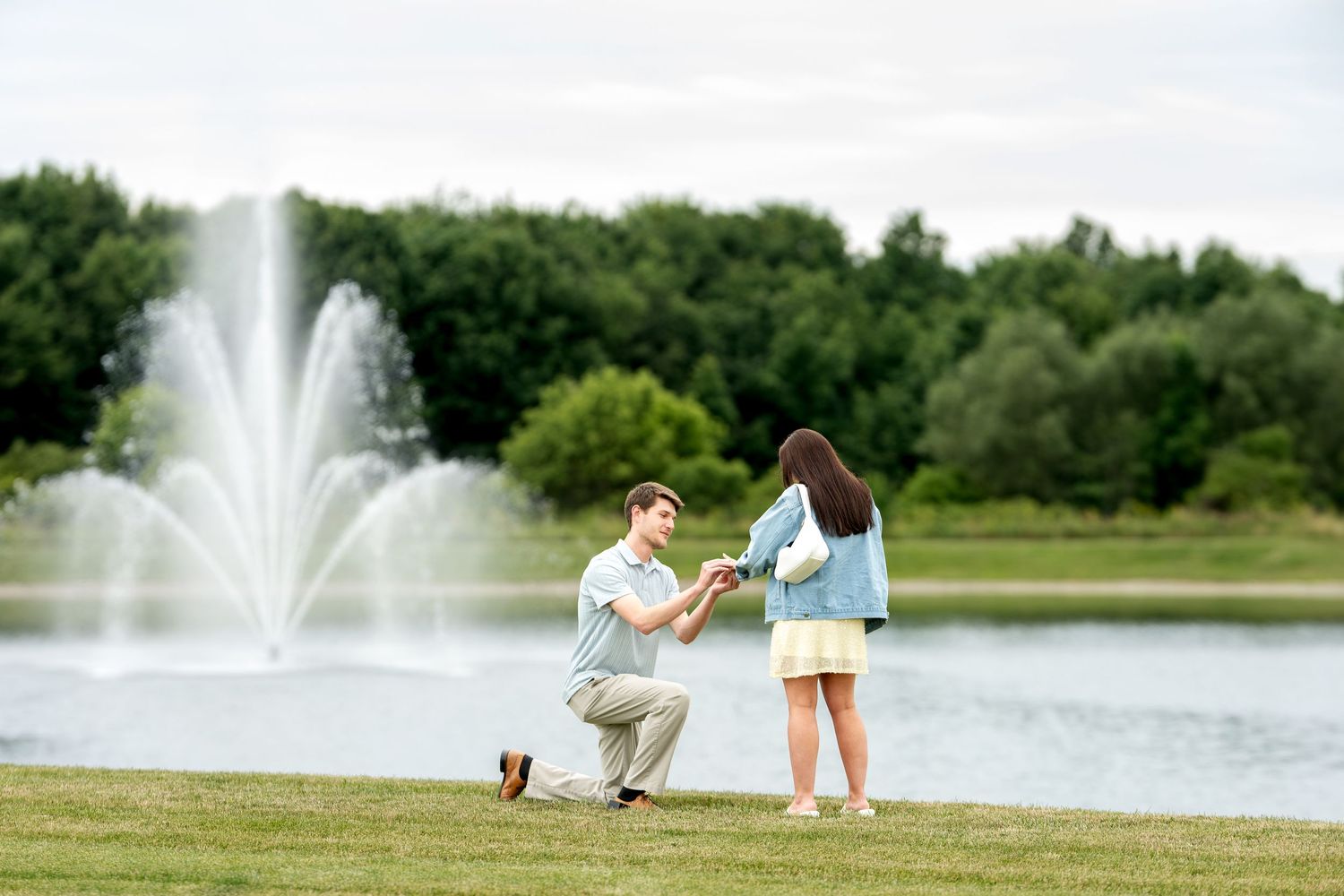Man proposes to woman on one knee by a scenic fountain in a lush green park on a sunny day.