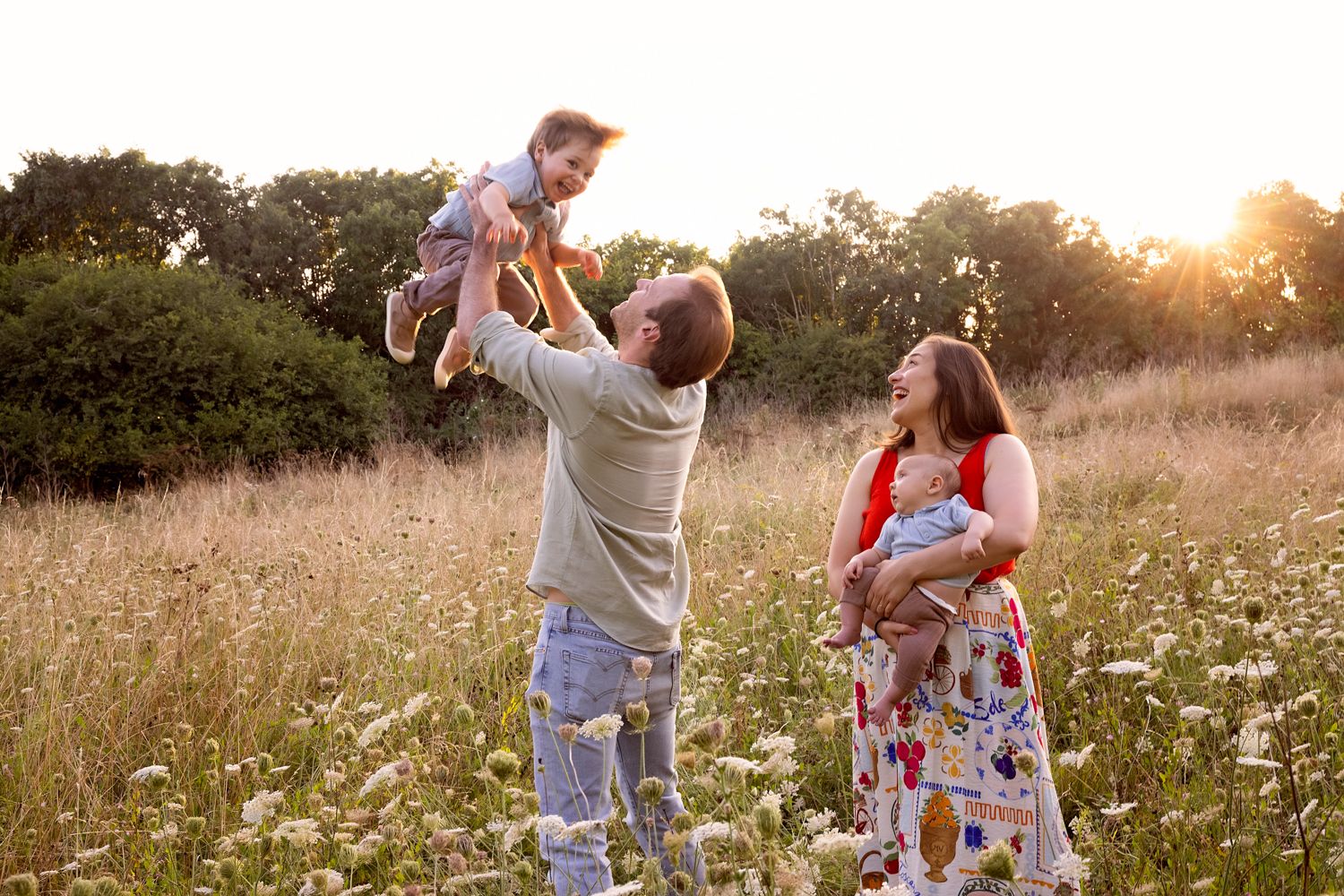 Family enjoying golden hour in a wildflower meadow, father lifting baby while mother watches and smiles.