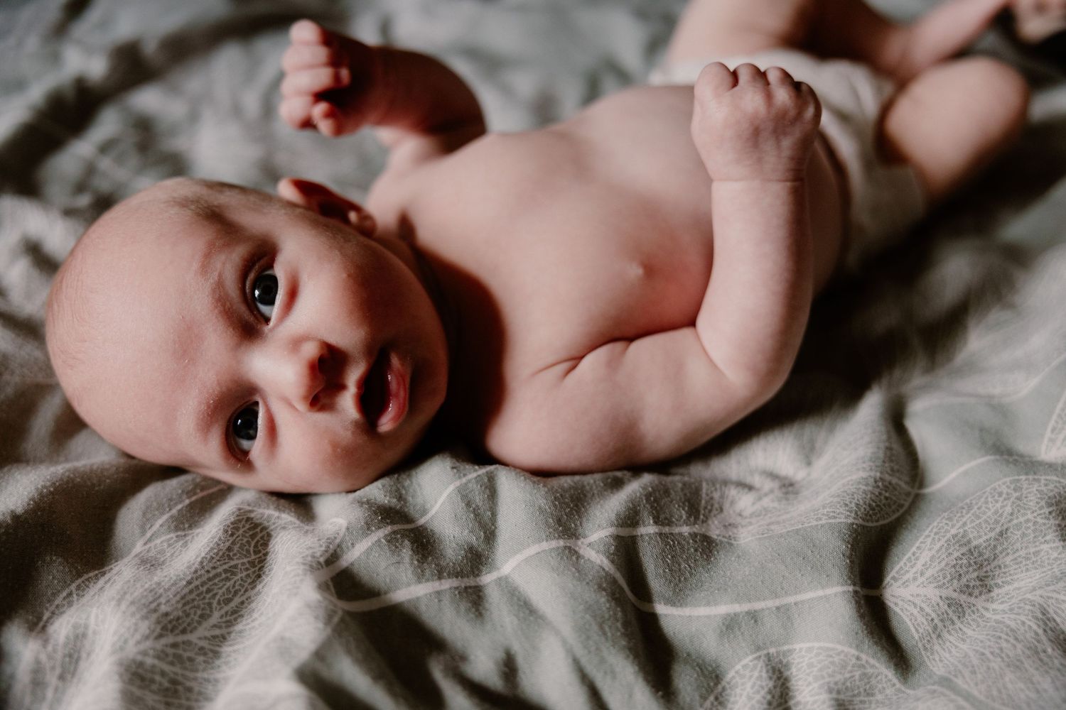 Newborn baby lying on a soft grey blanket, looking up with wide eyes and tiny arms raised.