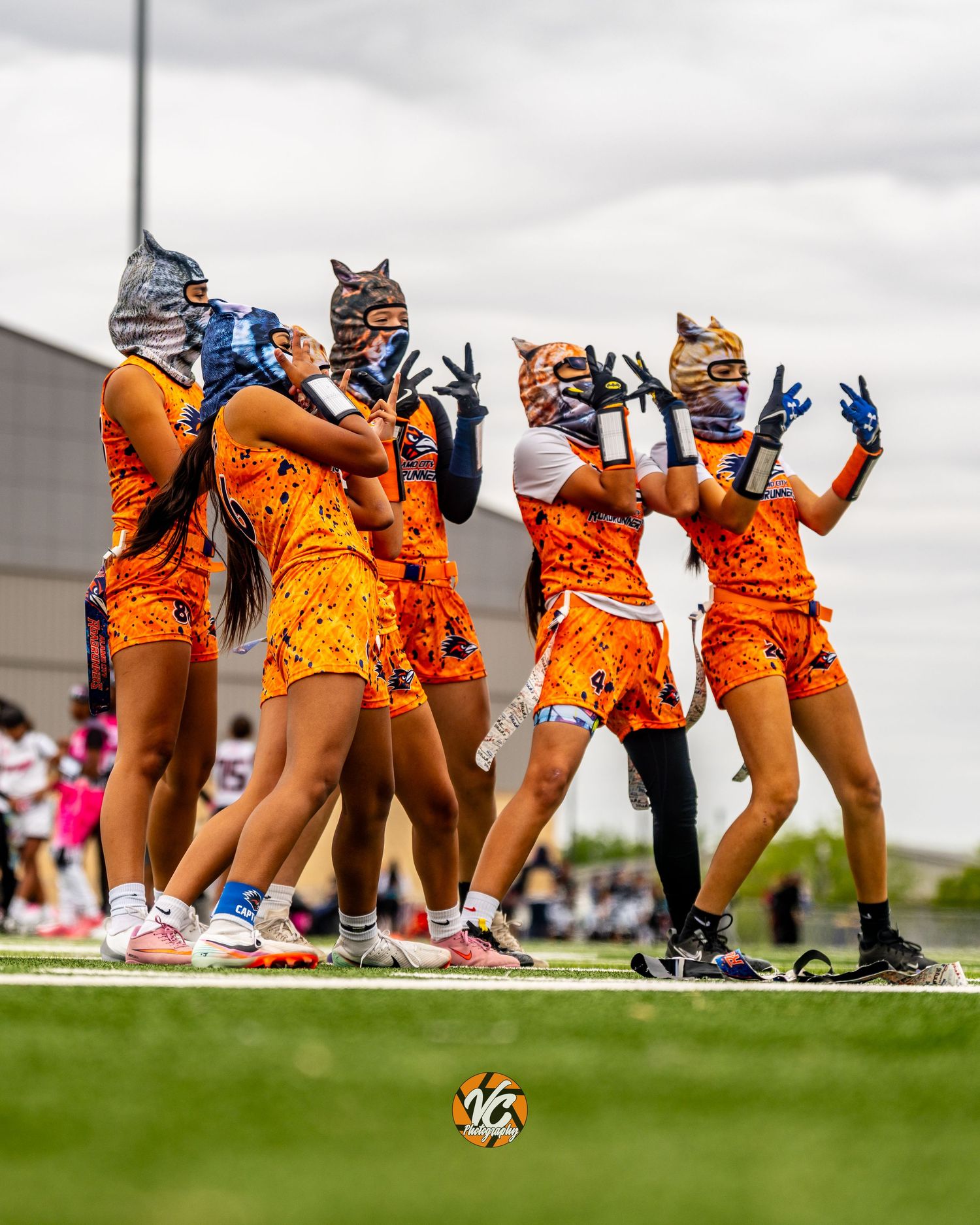 Girls in orange uniforms and cat masks celebrate on a green sports field during a competitive outdoor game.