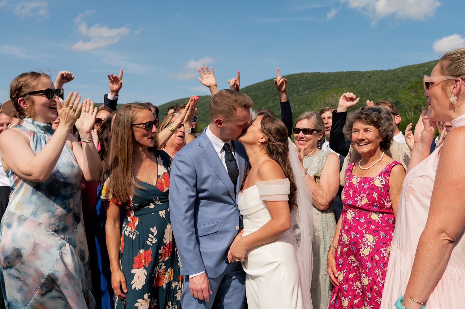 Wedding guests celebrate outdoors on a sunny day with mountains in the background and a couple standing in the center.