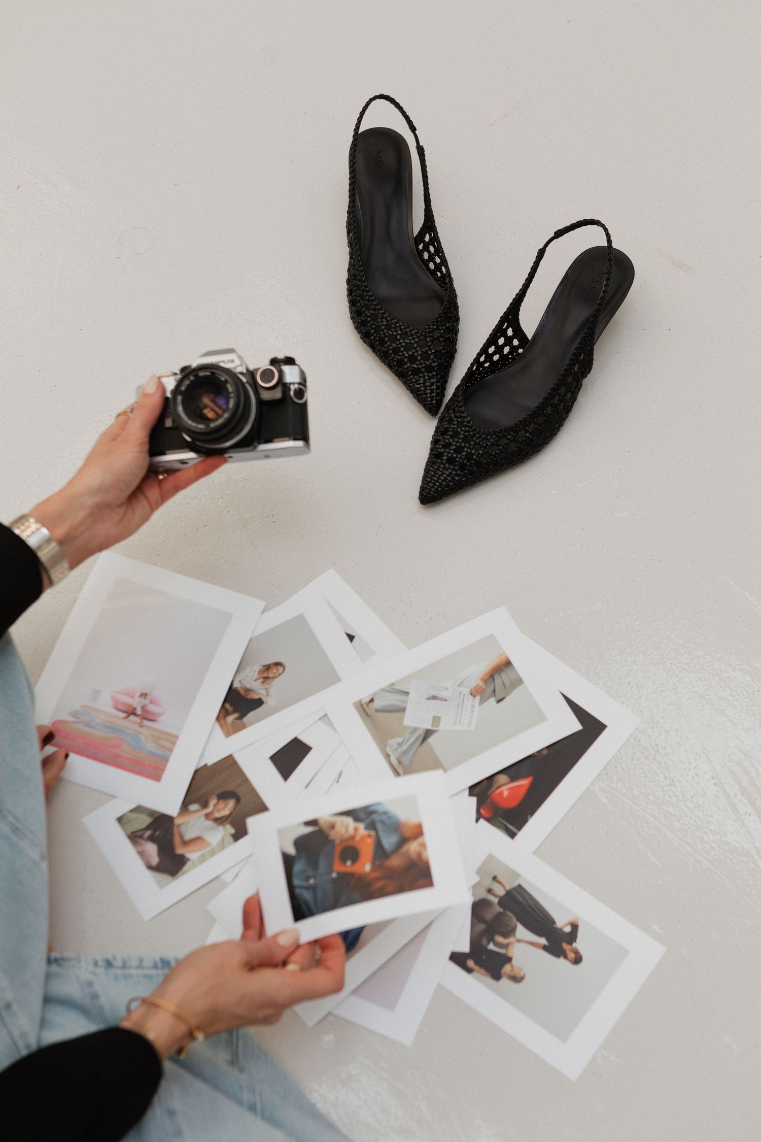 Hands holding vintage camera and photographs next to black lace slingback flats on white surface.