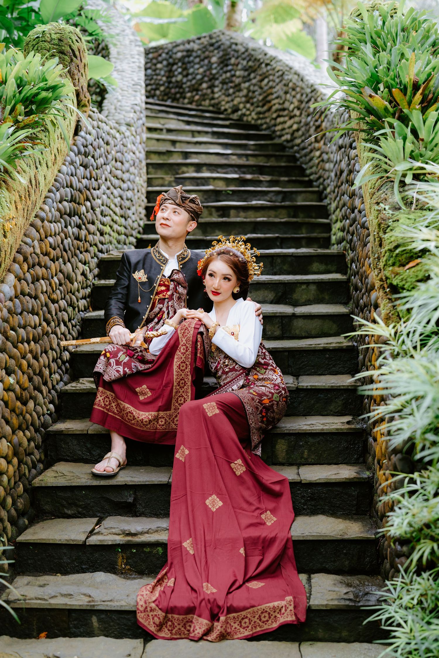 Couple in traditional Indonesian dress sits on stone steps with mosaic wall backdrop.