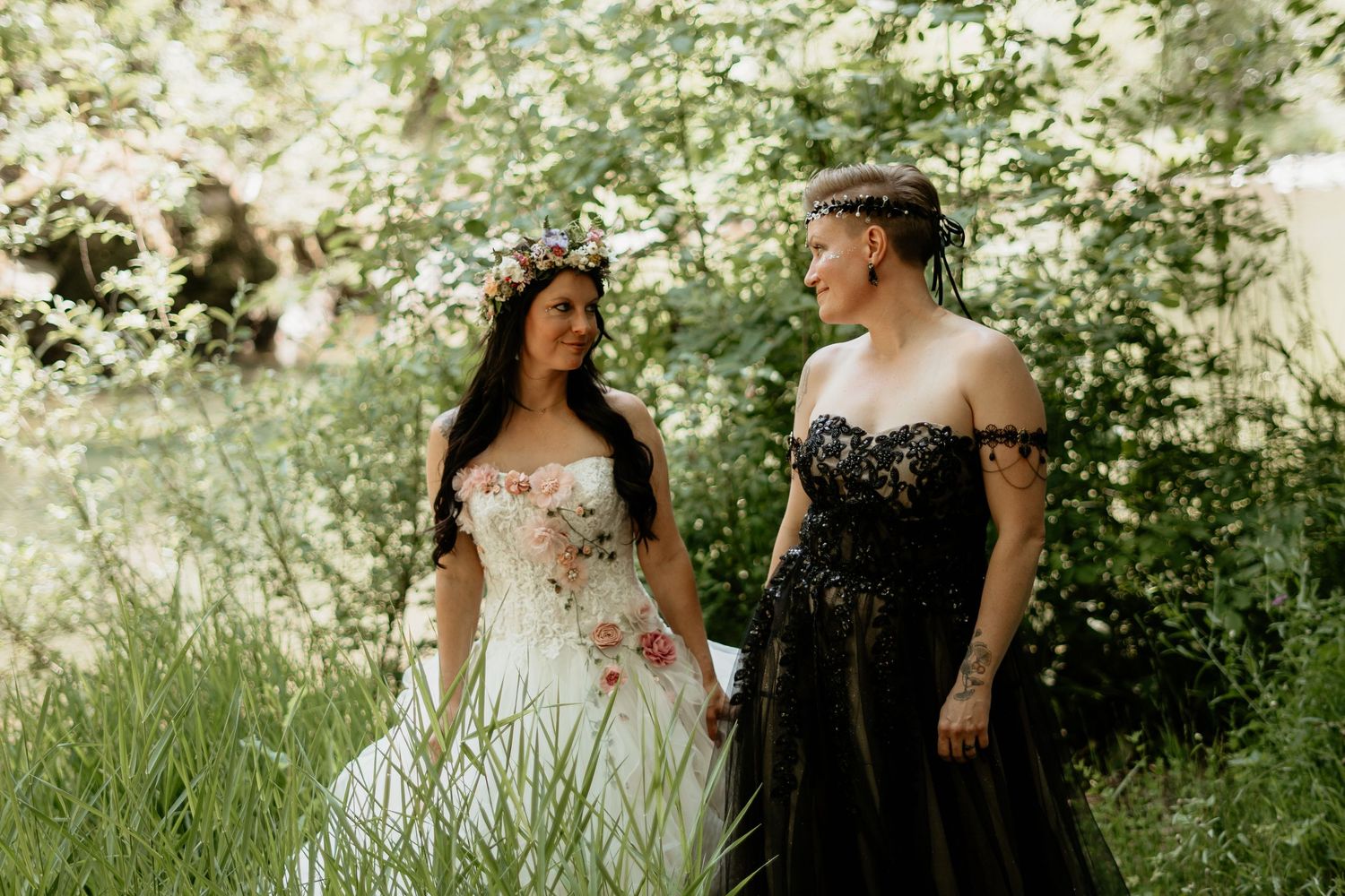 Two people in contrasting white and black formal dresses pose together in a sun-dappled garden setting with blossoming trees.