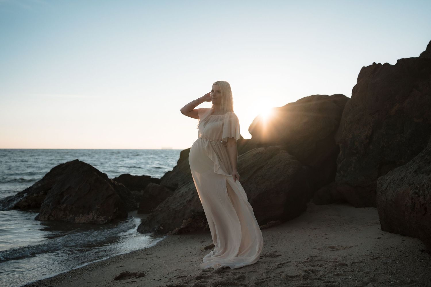 Silhouette of a person in flowing white dress standing on beach against rocky cliffs at sunset.