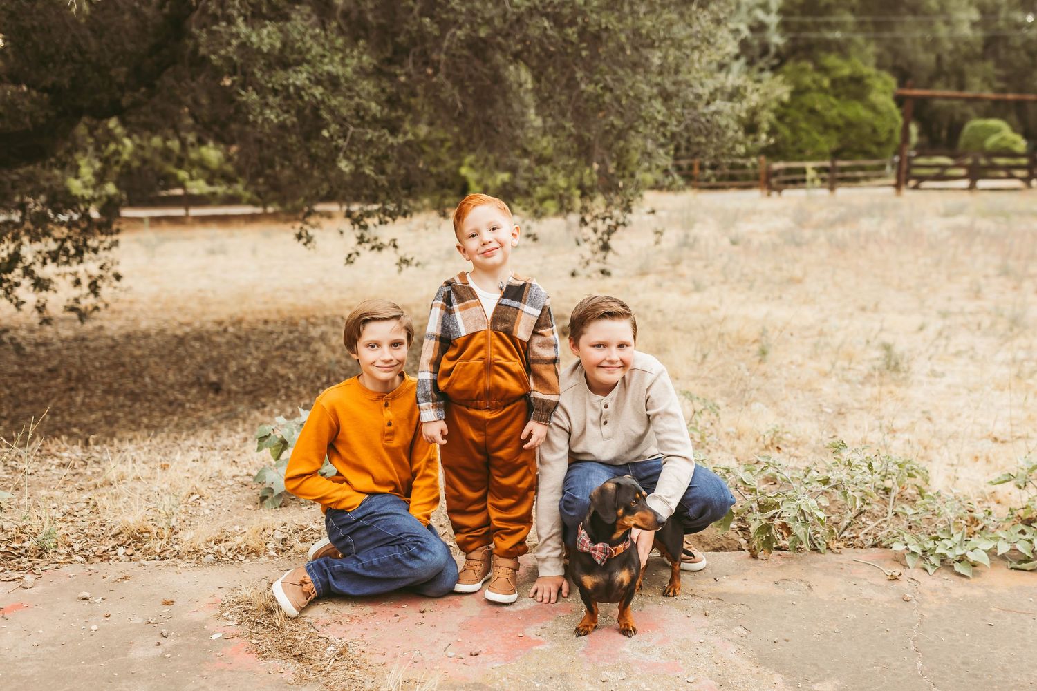 Three people and a small black dog sit together on a dirt path surrounded by trees in an outdoor setting.