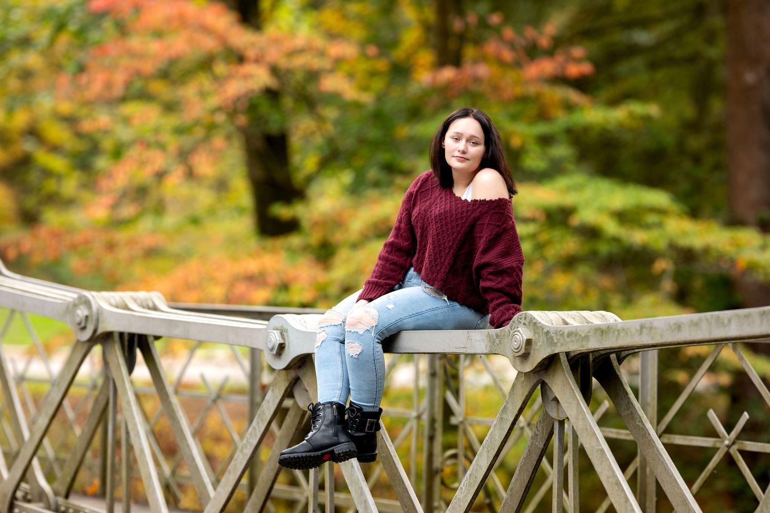 Teen girl in burgundy sweater sitting on wooden bridge surrounded by fall foliage.