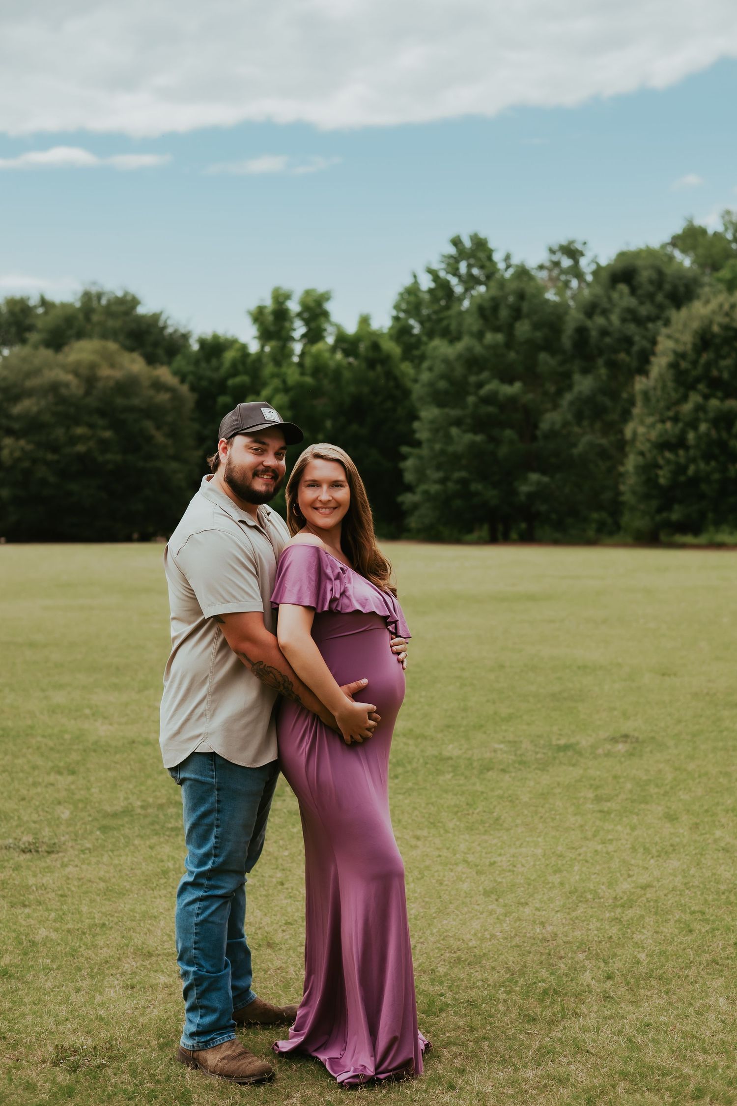 A couple stands together in a grassy field smiling and embracing while wearing casual attire during a Pinehurst, NC maternity photo shoot.