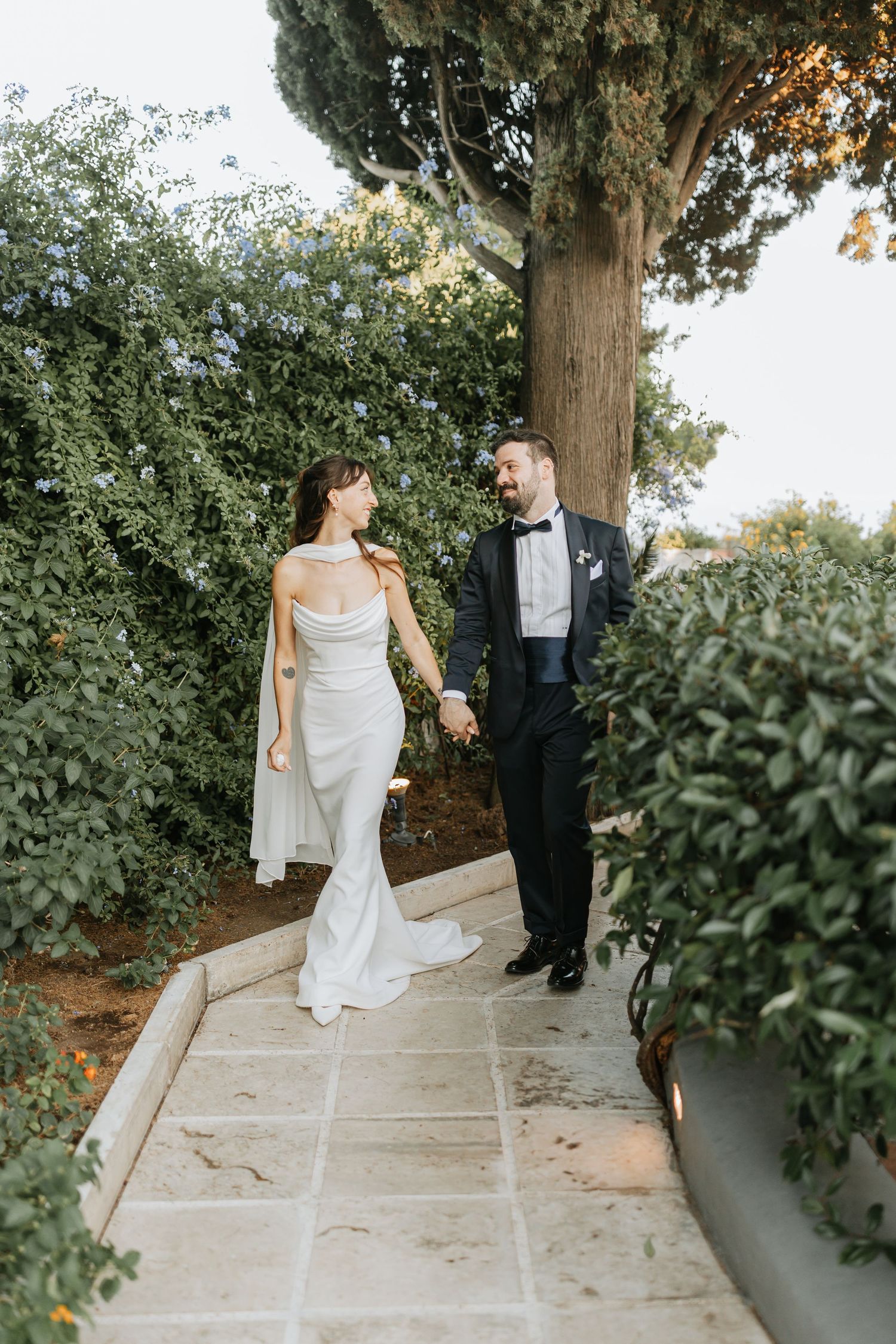 A couple in wedding attire walking along a stone pathway surrounded by lush greenery on their special day. Italy Wedding.