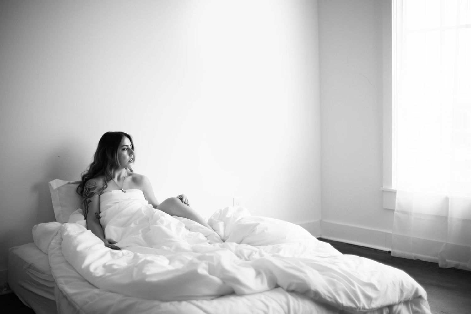 A person in a flowing white dress poses gracefully on a bed in a bright sunlit room.