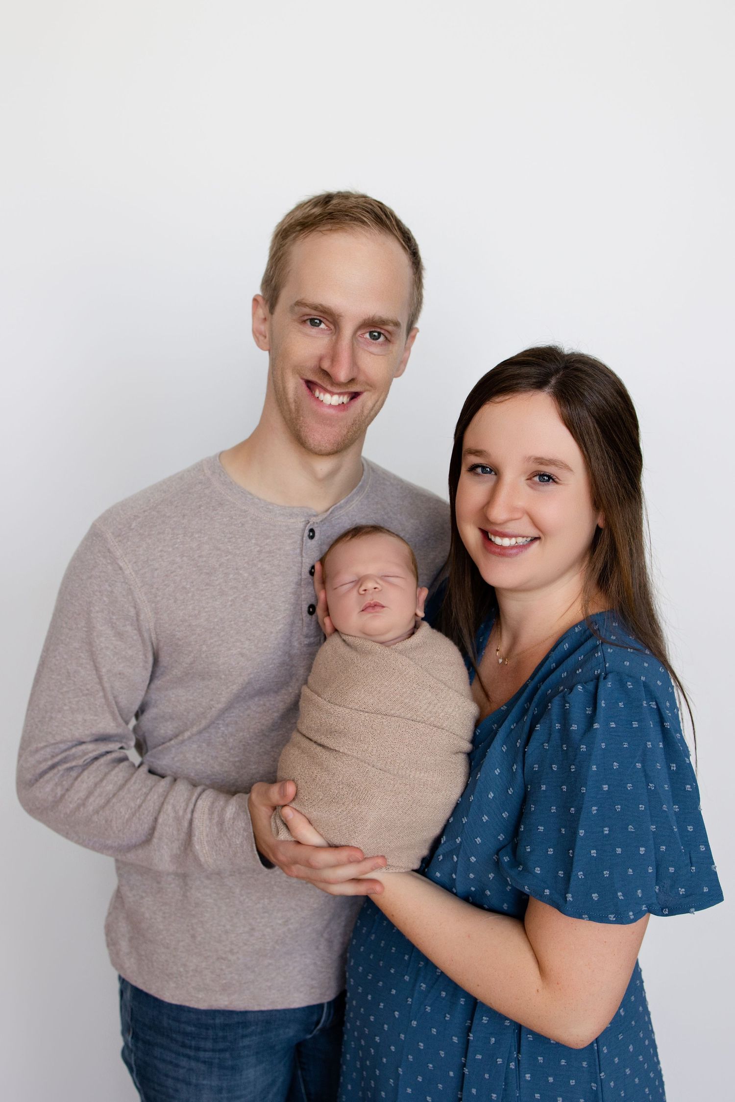 A couple in casual clothing holds their newborn baby wrapped in a beige swaddle against a white background.
