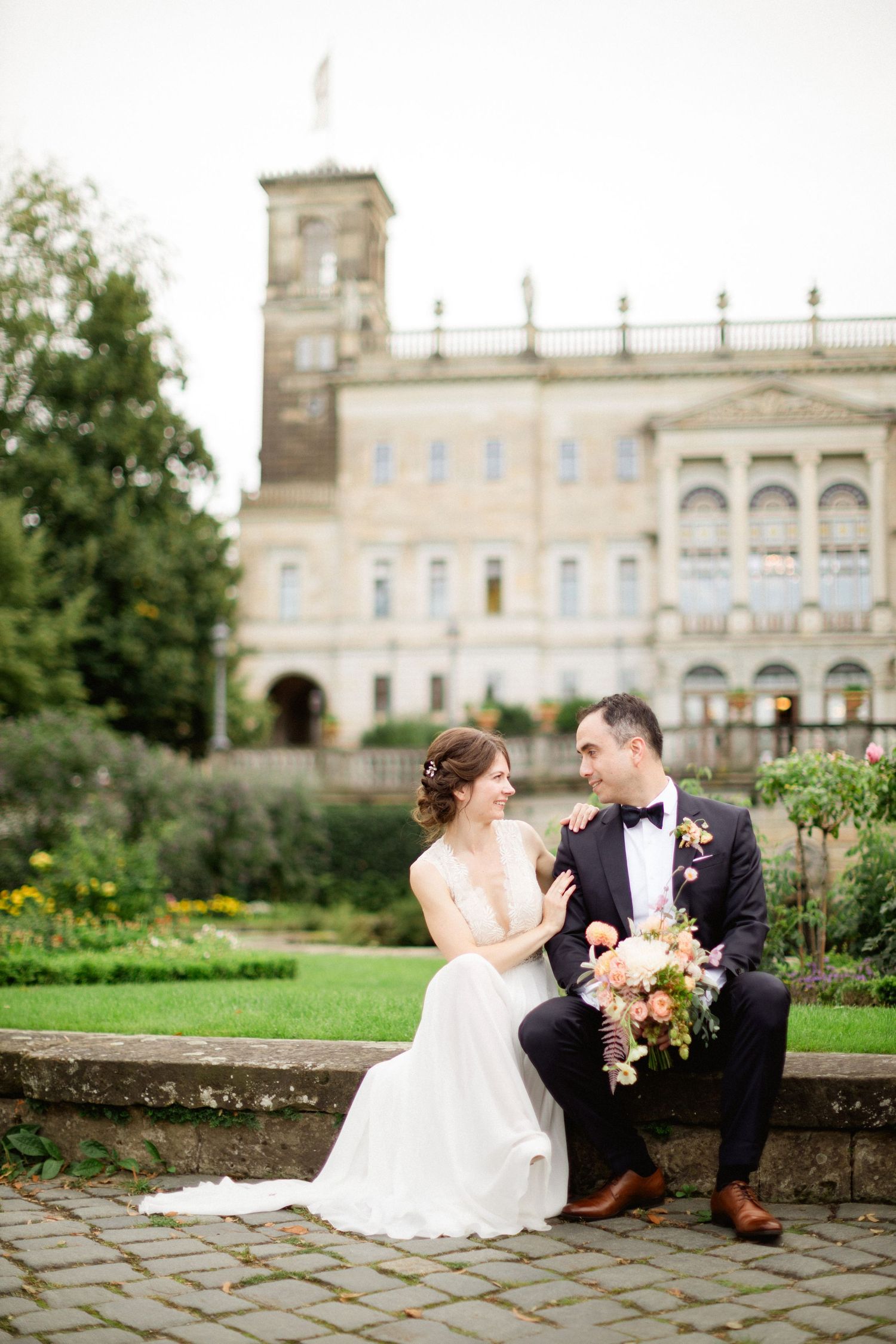Elegant wedding couple sitting on stone wall at historic estate with grandeur architecture and garden in background.