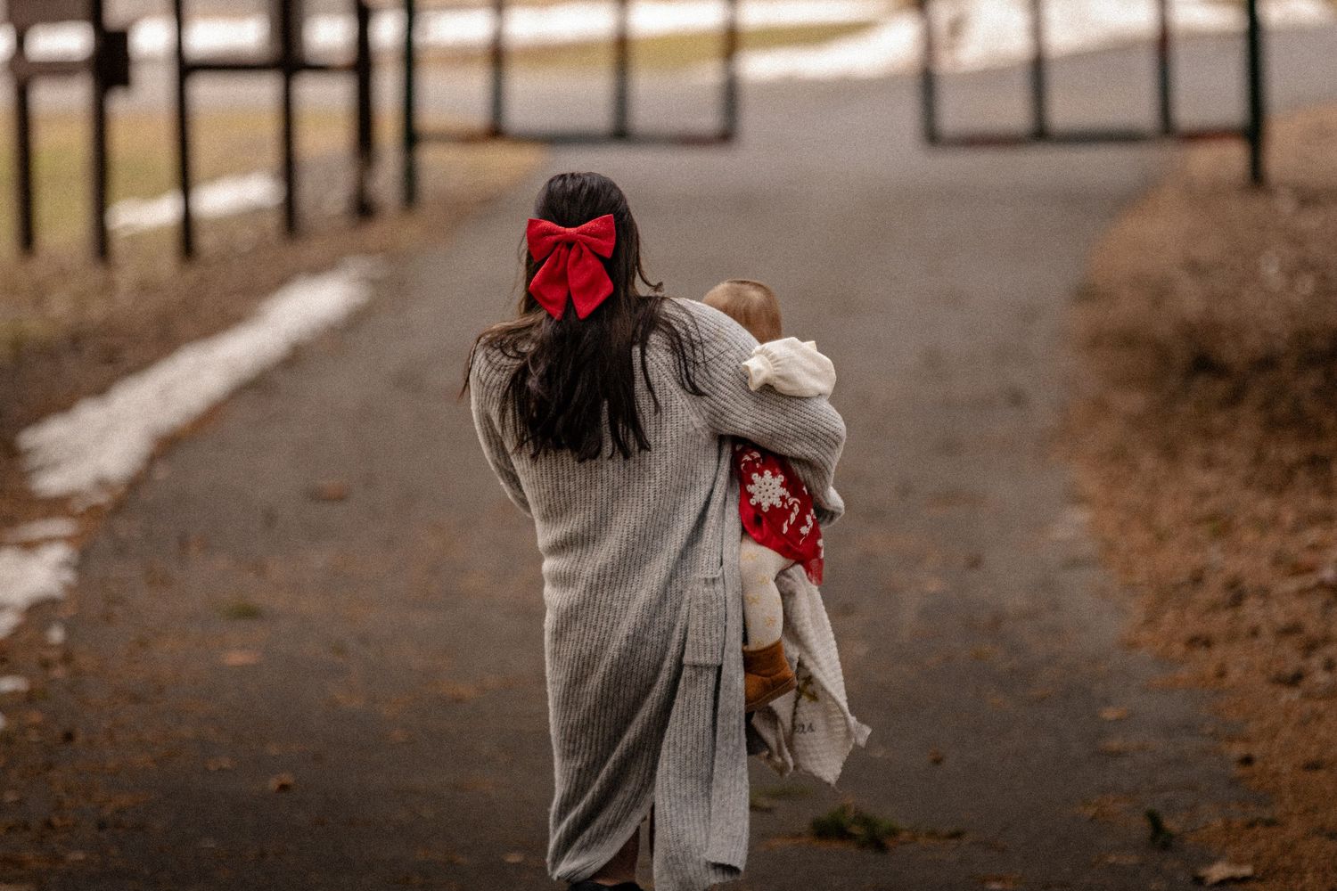 Someone wearing a gray coat walks down a tree-lined path while holding a stuffed animal on an autumn day.
