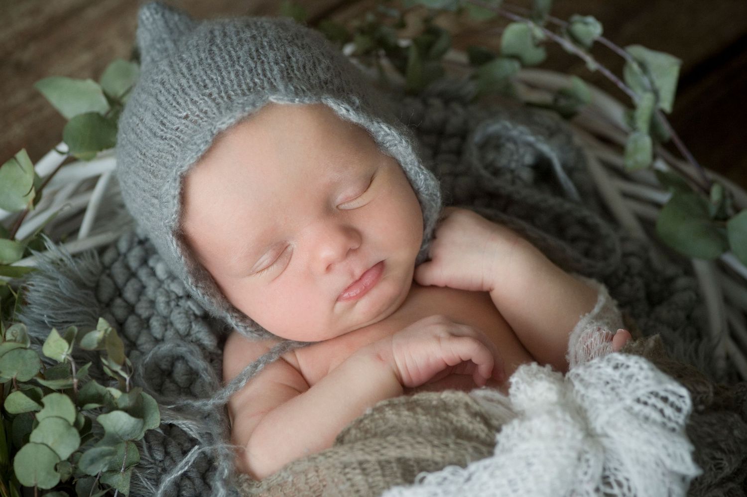 Sleeping newborn baby wearing a gray knit bonnet hat nestled in a rustic basket with green eucalyptus leaves.