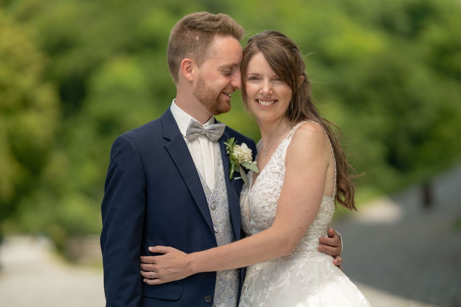A couple embraces while wearing wedding attire with a soft green natural background.
