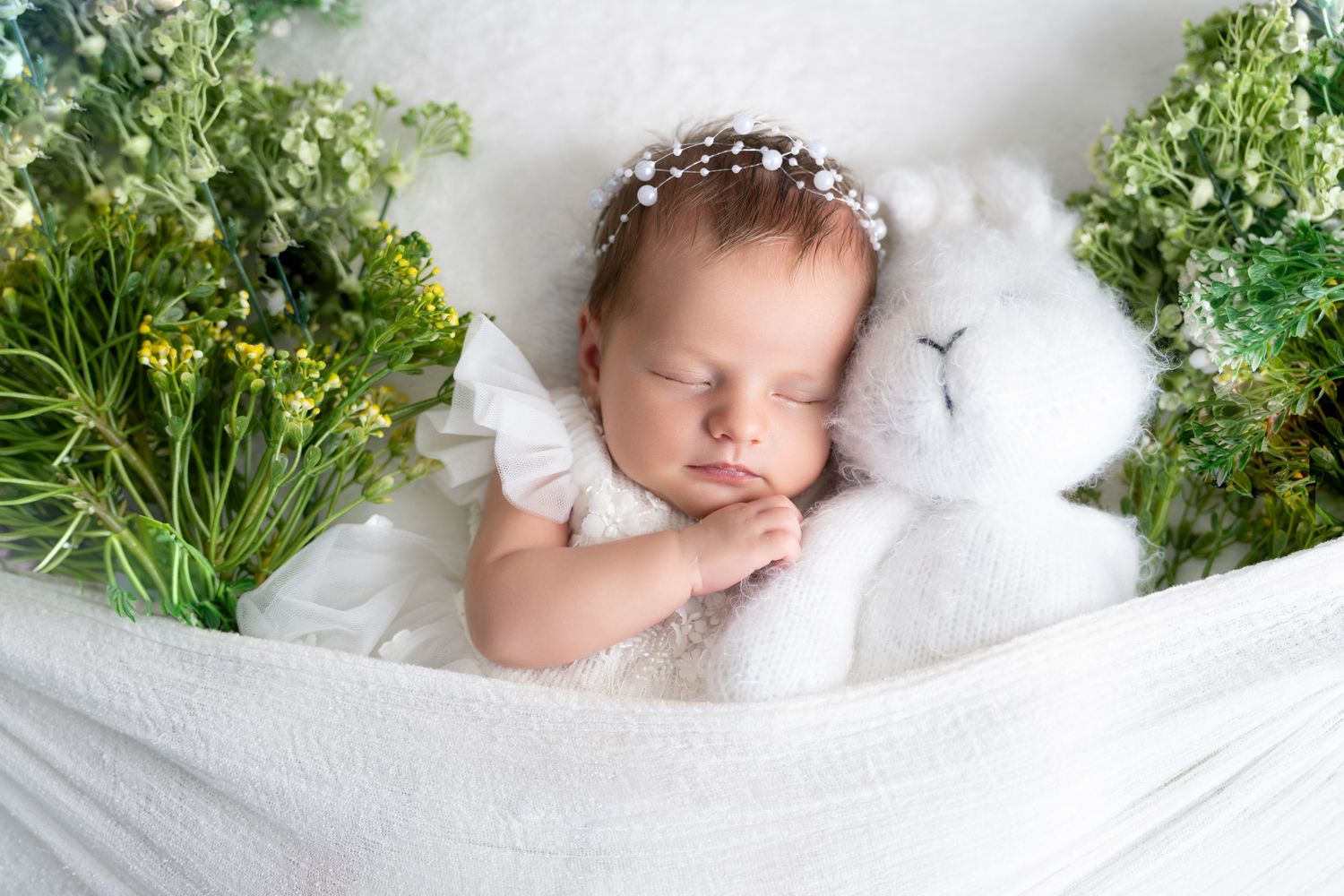 A sleeping baby nestled in soft white blankets surrounded by delicate green foliage and white flowers.