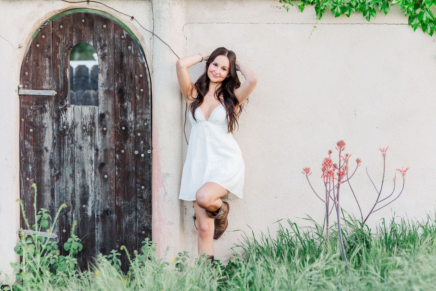 A woman in a white dress poses against a rustic wall with an old wooden door and green foliage.