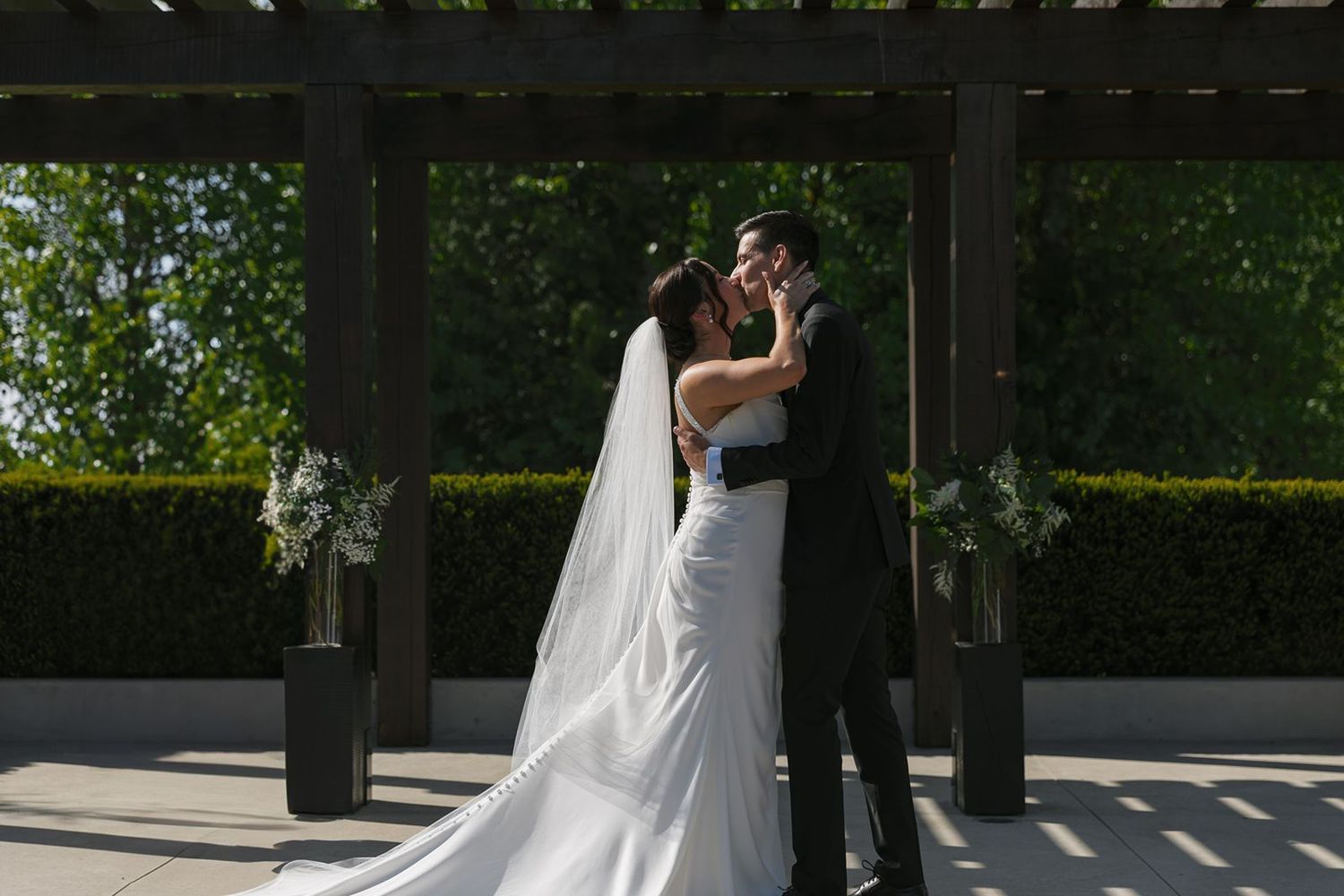 Bride and groom sharing a first kiss under a pergola surrounded by lush green hedges.