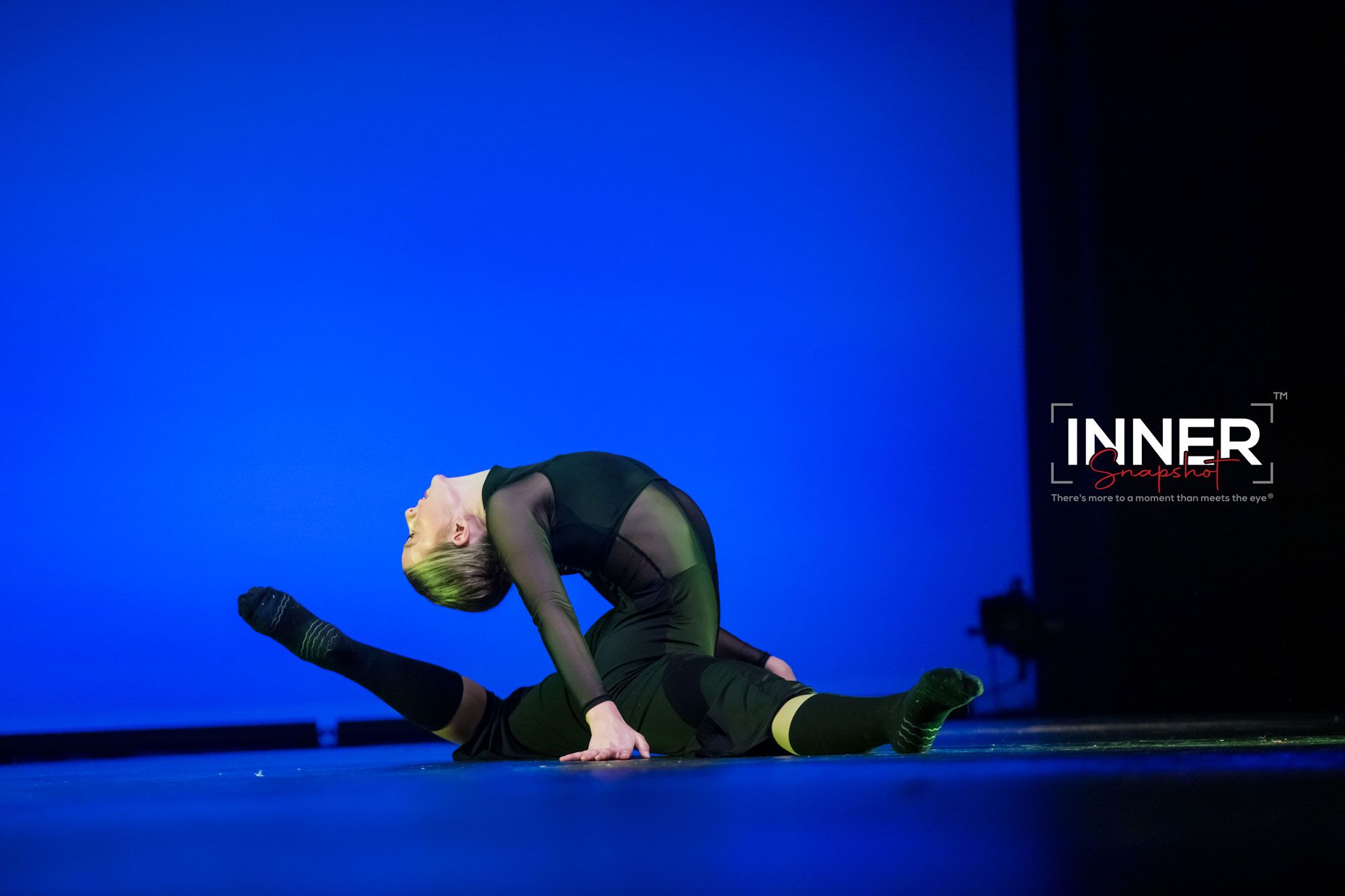 A performer, Niko Mulligan, in black clothing lies on a blue-lit stage floor during a dance performance, Poetry In Motion - Dec. 2025.