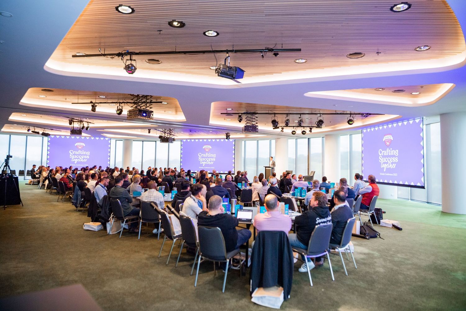 A panoramic view of a crowded modern conference room with blue lighting and rows of attendees seated at tables.
