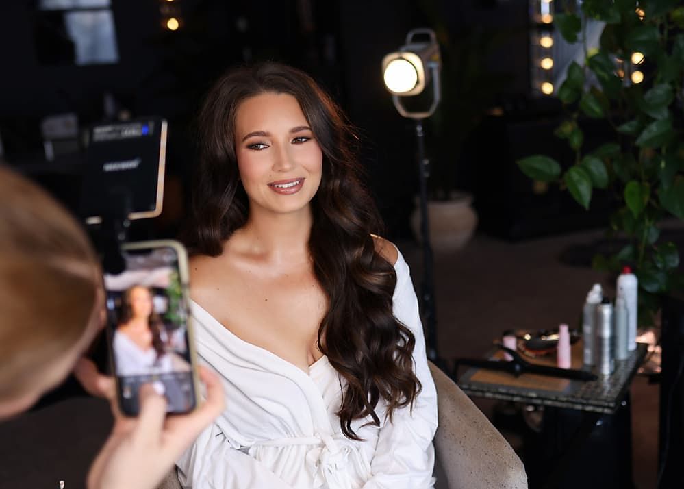 A woman in a white off-shoulder dress poses elegantly in front of a dark background with ambient lighting.