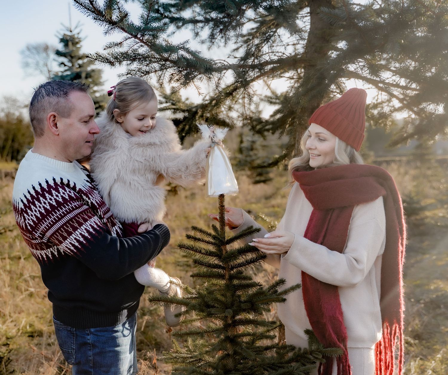Family in winter sweaters decorates a small evergreen Christmas tree together outdoors at sunset.