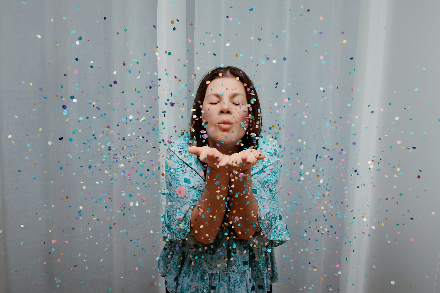 Standing against a grey backdrop, someone blows shimmering blue glitter from their palms while wearing a turquoise dress.